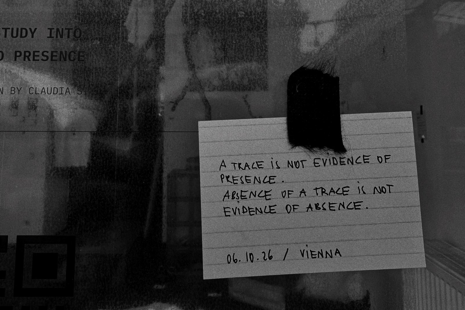 Handwritten index card fixed to the window with a tuft of dark fibre, reading: A trace is not evidence of presence. Absence of a trace is not evidence of absence. 06.10.26 / Vienna.